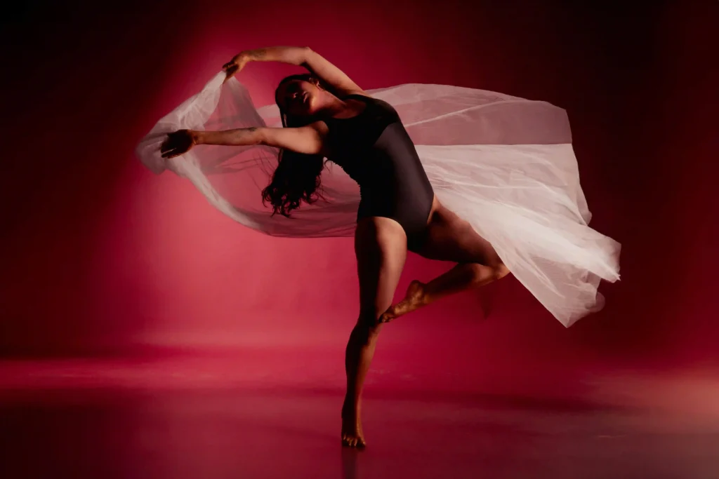 An artistic editorial portrait by a fashion stylist in Bangalore featuring a dancer in a black bodysuit with flowing white tulle against a dramatic red background.