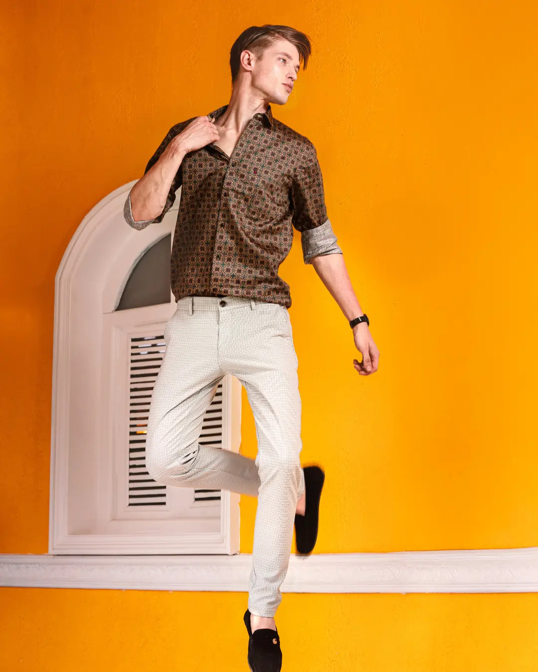 A male model captured in mid-air jumping against a bright yellow textured wall with a white arched window. Curated by a fashion stylist in Bangalore.