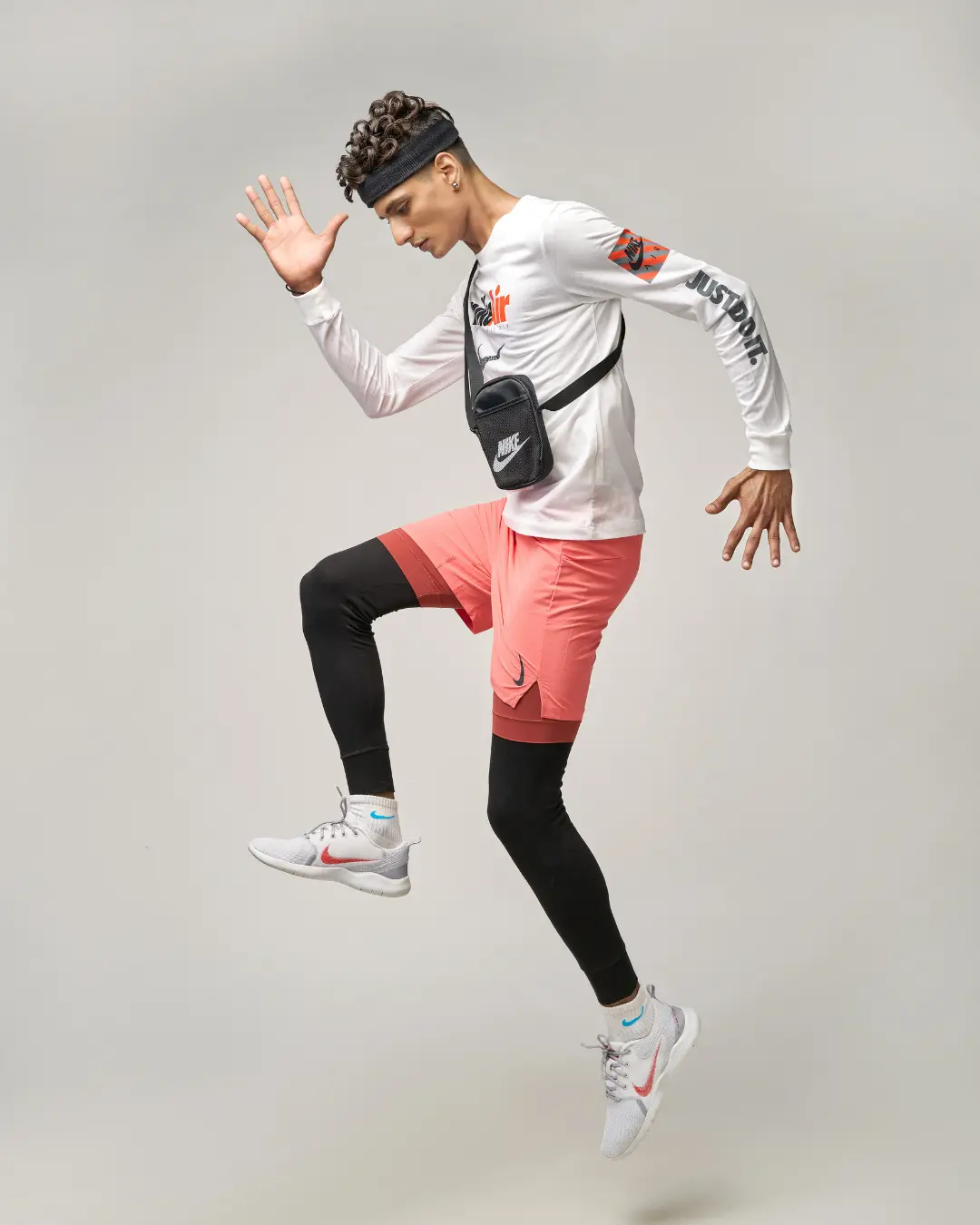 An action-packed studio shot curated by a fashion stylist in Bangalore, featuring a male model in mid-air wearing Nike athletic gear, including a white long-sleeve tee, coral shorts, and black leggings.
